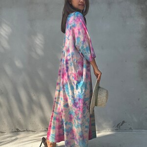 Op de afbeelding: Een vrouw draagt een lange, roze, blauwe en witte tie-dye jurk met een strohoed in haar hand. Ze staat voor een grijze betonnen muur.