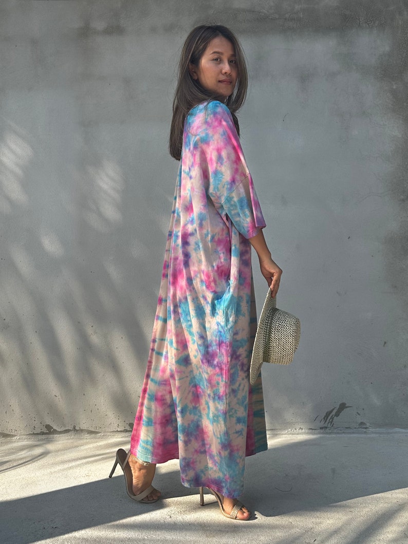 Op de afbeelding: Een vrouw draagt een lange, roze, blauwe en witte tie-dye jurk met een strohoed in haar hand. Ze staat voor een grijze betonnen muur.