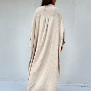 May include: A long, beige, oversized shirt dress with a collared neckline and long sleeves. The dress is made of a lightweight, textured fabric.