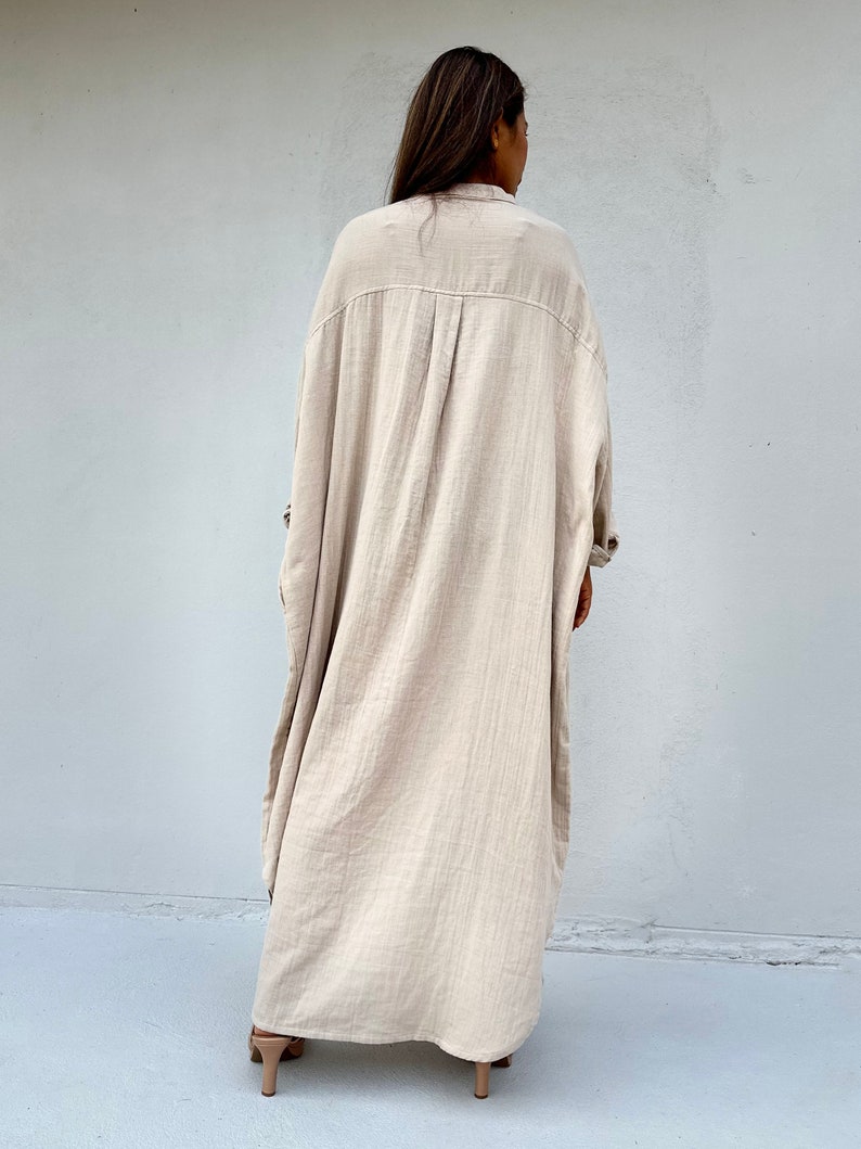 May include: A long, beige, oversized shirt dress with a collared neckline and long sleeves. The dress is made of a lightweight, textured fabric.