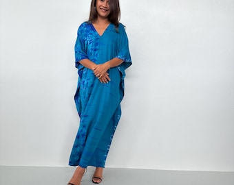Op de afbeelding: Een vrouw draagt een lange, vloeiende, blauwe tie-dye kaftan jurk. De jurk heeft een V-hals en wijde mouwen. De stof heeft een aquareleffect met blauwtinten. De vrouw draagt zwarte sandalen.