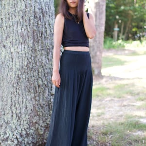 May include: A woman wearing a black crop top and black wide-leg pants stands in front of a tree.