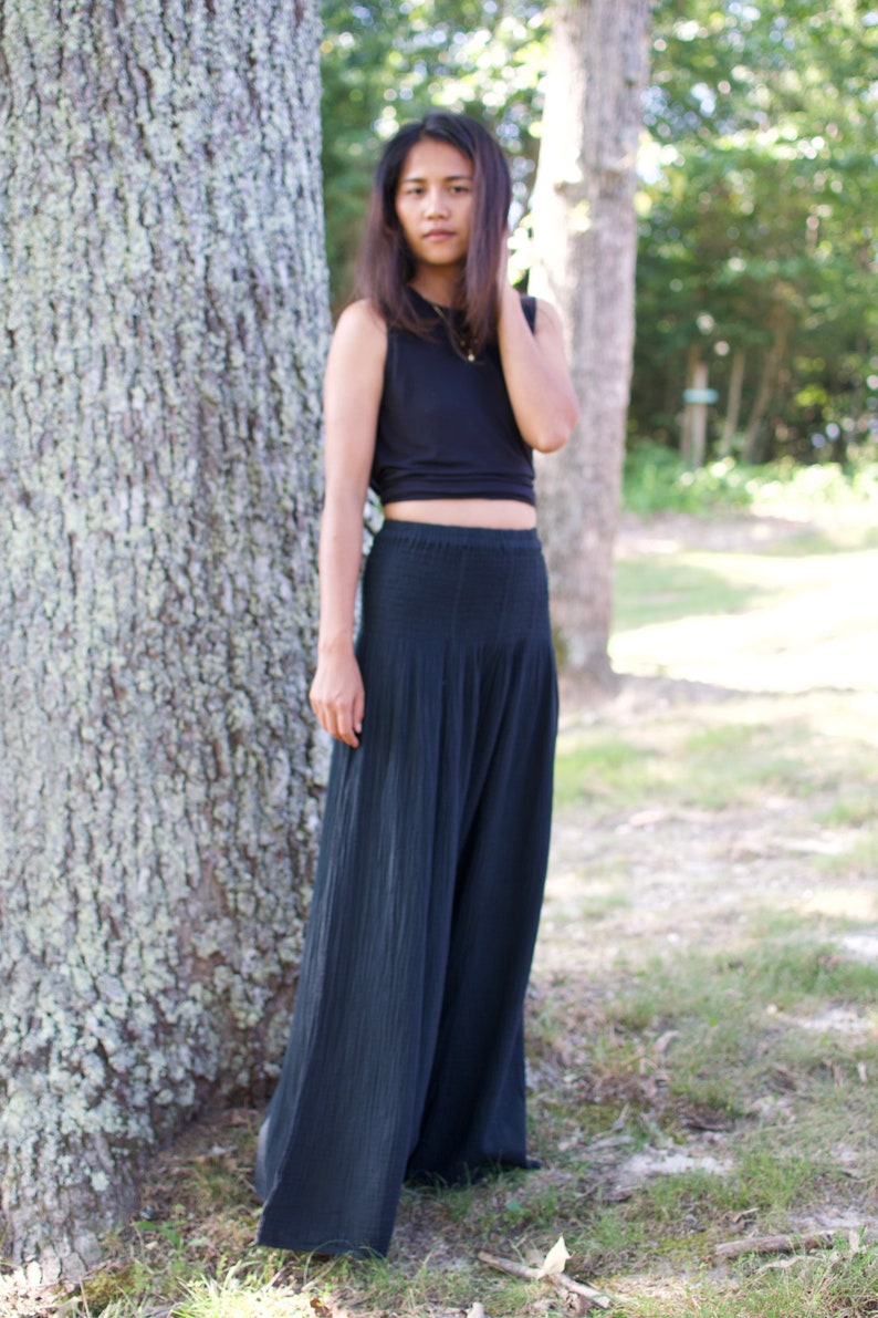 May include: A woman wearing a black crop top and black wide-leg pants stands in front of a tree.