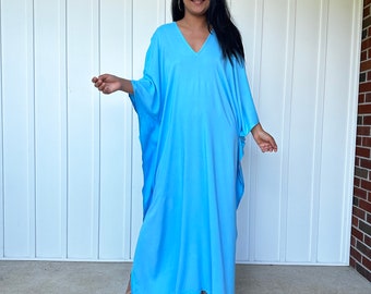 May include: A light blue, flowing caftan dress with a V-neck. The dress has wide sleeves and a loose fit. The model is wearing the dress and standing in front of a white wall. The dress is made of a soft, lightweight fabric.