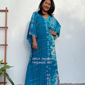 May include: A long, flowing blue and purple tie-dye kaftan with a V-neckline. The kaftan is made of a soft, lightweight fabric and has a relaxed fit. The text "MALA handworks Hollywood, MD" is printed on the front of the kaftan.
