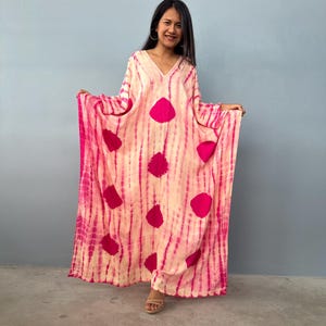 May include: A long, flowing, tie-dye dress in shades of pink and cream. The dress features a V-neck and large, circular pink accents. The fabric has vertical stripes of pink and cream. The dress is worn by a person.