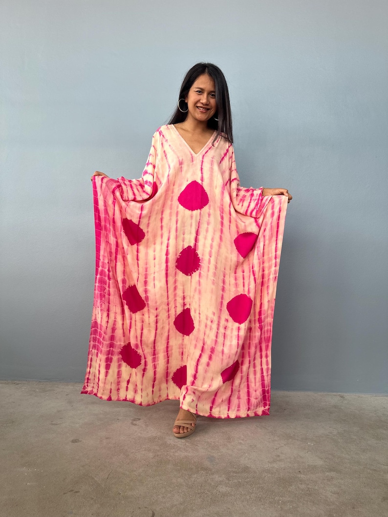 May include: A long, flowing, tie-dye dress in shades of pink and cream. The dress features a V-neck and large, circular pink accents. The fabric has vertical stripes of pink and cream. The dress is worn by a person.