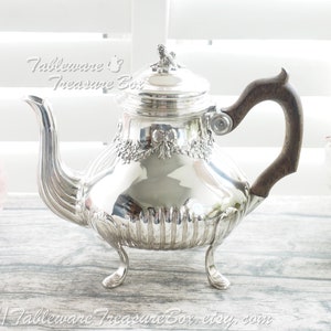 May include: A silver teapot with a wooden handle and three legs. The teapot has a decorative design around the top and a fluted body.