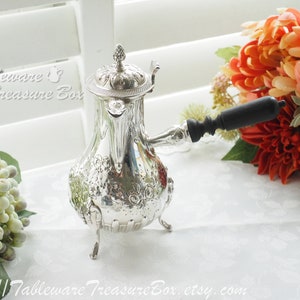 May include: A silver coffee pot with an ornate design and a black wooden handle. The pot is sitting on a white tablecloth with a floral pattern.