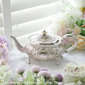 May include: An ornate silver teapot with detailed floral and scroll patterns. The teapot has a curved spout, a loop handle, and a decorative lid. It is displayed on a white surface, surrounded by flowers. The text "Tableware Treasure Box" is visible.