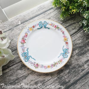 May include: A white porcelain dinner plate with a gold rim and a floral design featuring blue bows and pink, yellow, and green flowers.