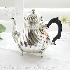 May include: A silver teapot with a black handle and a swirling design. The teapot has three feet and a lid with a finial. The teapot is on a white tablecloth.