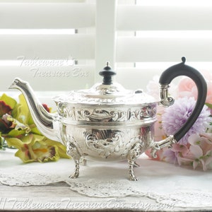 May include: A silver teapot with ornate detailing and a black handle. The teapot has three legs and is sitting on a white lace tablecloth.