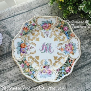 May include: A white porcelain plate with a gold and floral design. The plate has a scalloped edge and features a central floral motif with gold accents. The plate is decorated with a variety of flowers, including roses, lilies, and daisies.