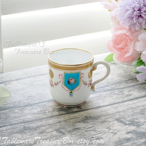 May include: A white porcelain teacup with a gold rim and handle, featuring a blue and pink floral crest design. The cup has a gold band around the top and is decorated with pink floral accents. The background includes flowers and the text "Tableware's Treasure Box".