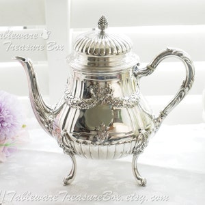 May include: A silver teapot with a fluted lid and ornate details. The teapot has three legs and a curved spout. The teapot is on a white surface.