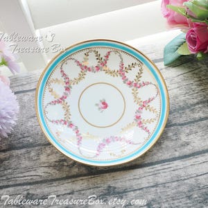 May include: A white porcelain saucer with a gold rim and a light blue border. The saucer is decorated with pink floral garlands and a small pink rose in the centre. The text "Tableware's Treasure Box" and a website address are visible.