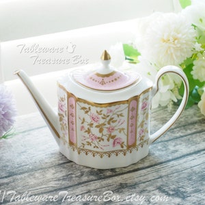 May include: A white teapot with a pink and gold floral design. The teapot has a lid with a pink and gold trim. The teapot is sitting on a wooden surface.