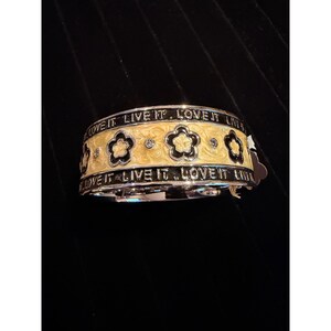 May include: A silver-toned bracelet with a yellow enamel band featuring black flower designs and the text "LOVE IT LIVE IT." The bracelet has a repeating floral pattern and small, sparkling accents.
