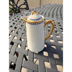 May include: A white porcelain coffee pot featuring a cylindrical body, a gold-toned handle, and a matching lid. The rim of the lid and the upper section of the pot are decorated with a delicate blue leaf pattern and gold trim accents.