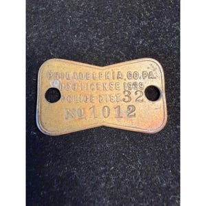May include: A brass-colored metal dog license tag from Philadelphia, PA, dated 1939. The rectangular tag has a notched center, two circular holes on the sides, and embossed text reading PHILADELPHIA CO. PA. DOG LICENSE 1939 POLICE DIST. 32 NO. 1012.