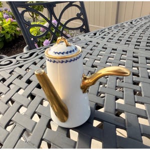 May include: A white porcelain coffee pot featuring a gold-toned handle and spout. The cylindrical body and lid are decorated with a delicate blue leaf pattern and gold trim. The lid has a small gold finial at the top center.