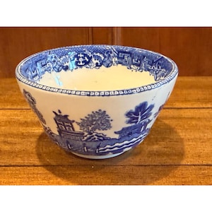 May include: A ceramic bowl featuring a blue and white transferware pattern. The exterior displays a detailed landscape with pagodas, trees, and water elements. The interior rim is decorated with a geometric border pattern in blue and white.