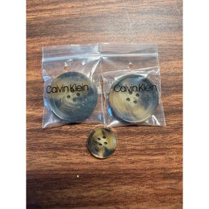 May include: Two round, brown, four-hole buttons, each individually sealed in a clear plastic bag labeled with the text Calvin Klein, accompanied by one loose, matching brown four-hole button positioned below the two plastic bags.