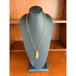 May include: A gold-toned necklace featuring a long, slender chain and a geometric pendant. The pendant consists of a rectangular top section with four vertical, dangling bars of varying lengths, creating a tiered, fringe-like appearance.