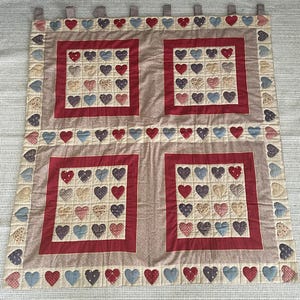 May include: A rectangular quilted wall hanging featuring four square panels, each containing a grid of sixteen small fabric hearts in shades of red, blue, and cream. The border is decorated with a continuous row of hearts and fabric tabs.