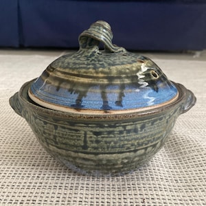May include: A round ceramic lidded casserole dish featuring a mottled olive green and blue glaze. The lid includes a sculpted handle and small steam vents. The base has two small side handles and a textured, speckled finish throughout.