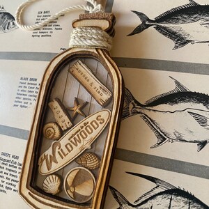 May include: A wooden bottle ornament with a cork top and a clear glass insert. The glass insert contains a small surfboard with the text "Welcome to the Wildwoods" and other nautical themed decorations. The bottle is decorated with a gold rope and the text "Amanda & Stan 2023".