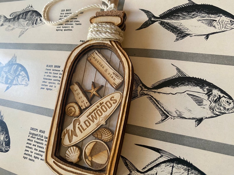 May include: A wooden bottle ornament with a cork top and a clear glass insert. The glass insert contains a small surfboard with the text "Welcome to the Wildwoods" and other nautical themed decorations. The bottle is decorated with a gold rope and the text "Amanda & Stan 2023".