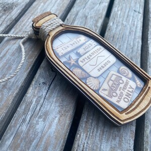 May include: A wooden ornament shaped like a bottle with a cork top and a white twine handle. The bottle contains a variety of small wooden cutouts, including seashells, a starfish, a 4x4 truck, and a sign that reads "the best briganttine season 2023" and another sign that reads "NJDEP Permit Only".