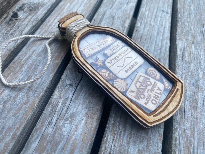 May include: A wooden ornament shaped like a bottle with a cork top and a white twine handle. The bottle contains a variety of small wooden cutouts, including seashells, a starfish, a 4x4 truck, and a sign that reads "the best briganttine season 2023" and another sign that reads "NJDEP Permit Only".