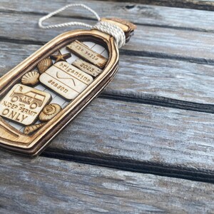 May include: Wooden ornament shaped like a bottle with a cork stopper. The bottle contains a variety of small wooden cutouts, including seashells, a starfish, a 4x4 truck, and a sign that reads "4x4 with NUDE PERMIT ONLY". The bottle also has a small wooden sign that reads "The Bells 2023 Brigantine Season".
