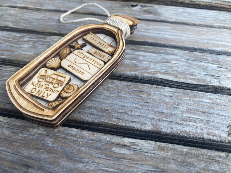 May include: Wooden ornament shaped like a bottle with a cork stopper. The bottle contains a variety of small wooden cutouts, including seashells, a starfish, a 4x4 truck, and a sign that reads "4x4 with NUDE PERMIT ONLY". The bottle also has a small wooden sign that reads "The Bells 2023 Brigantine Season".