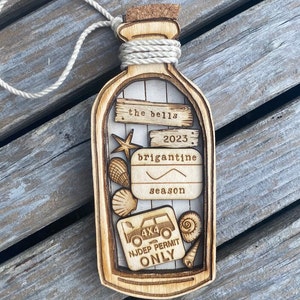 May include: A wooden ornament shaped like a bottle with a cork top and a rope tied around the neck. Inside the bottle are various wooden cutouts including seashells, a starfish, a car, and a sign that reads "the bells 2023 brigantine season 4x4 with NJDEP PERMIT ONLY".