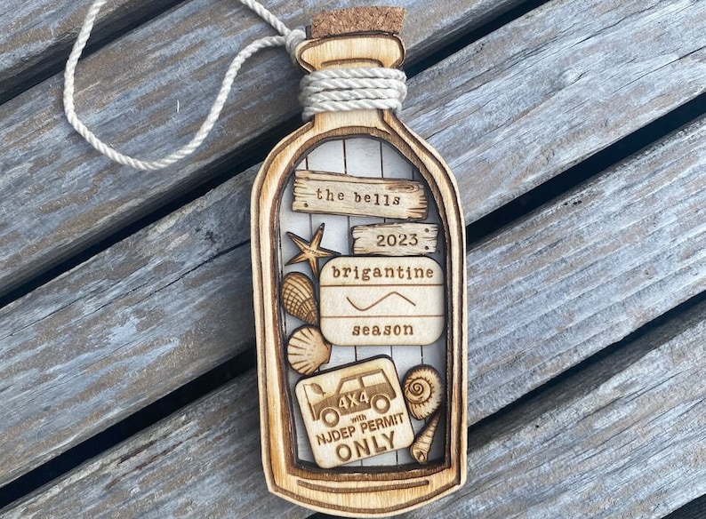 May include: A wooden ornament shaped like a bottle with a cork top and a rope tied around the neck. Inside the bottle are various wooden cutouts including seashells, a starfish, a car, and a sign that reads "the bells 2023 brigantine season 4x4 with NJDEP PERMIT ONLY".