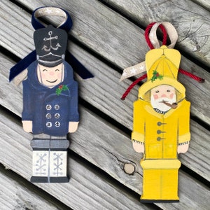 May include: Two wooden Christmas ornaments, one depicting a sailor wearing a yellow raincoat and holding a pipe, and the other depicting a sailor wearing a blue coat and a hat with an anchor on it.