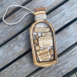 May include: A wooden ornament shaped like a bottle with a cork top and a rope tied around the neck. The bottle contains a variety of small wooden cutouts, including seashells, a starfish, a fishing hook, and a 4x4 vehicle with the text "NJDEP PERMIT ONLY". The bottle also has the text "the bells 2023 brigantine season" on it.
