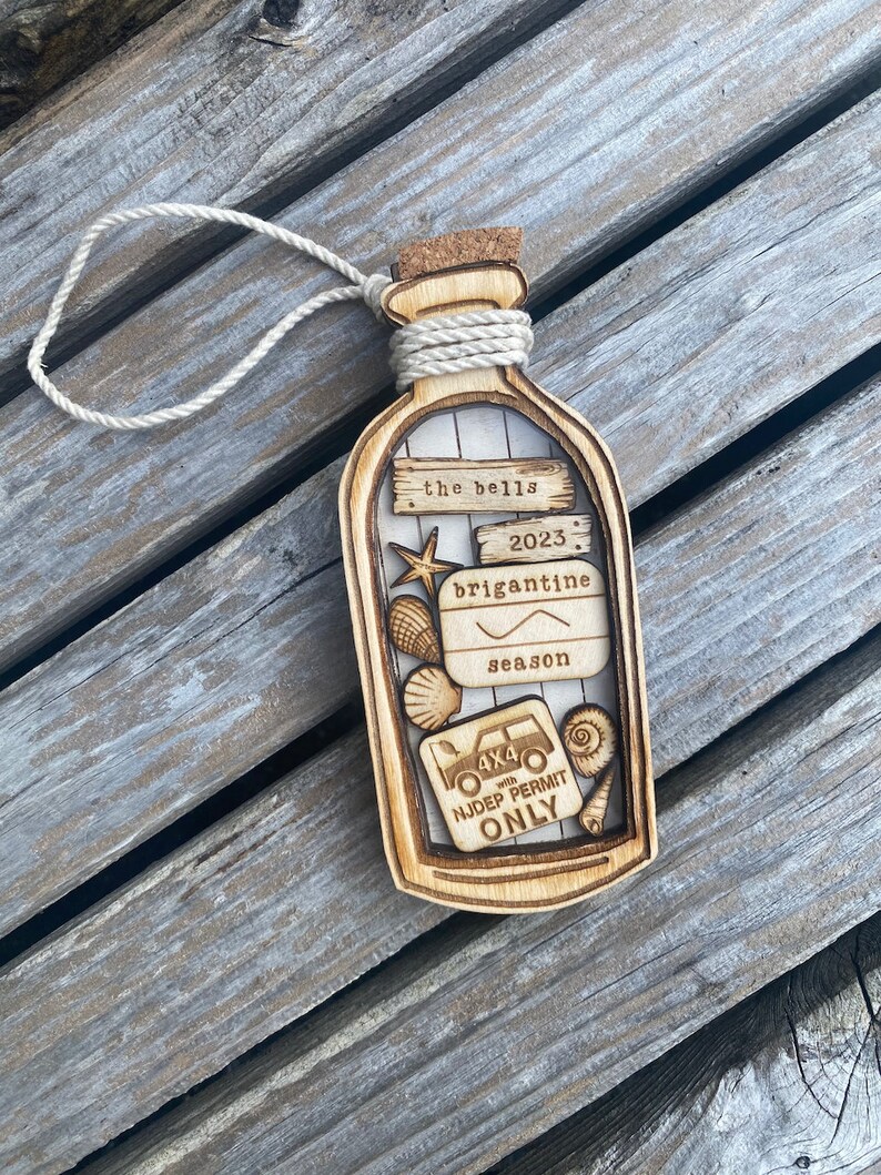 May include: A wooden ornament shaped like a bottle with a cork top and a rope tied around the neck. The bottle contains a variety of small wooden cutouts, including seashells, a starfish, a fishing hook, and a 4x4 vehicle with the text "NJDEP PERMIT ONLY". The bottle also has the text "the bells 2023 brigantine season" on it.