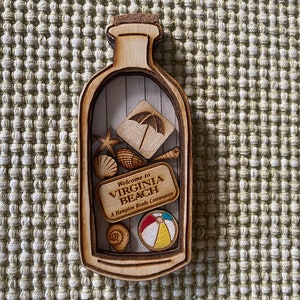 May include: Wooden bottle-shaped ornament with a cork top. Inside the bottle are various wooden cutouts including a beach umbrella, seashells, a starfish, a seashell, a beach ball, and a sign that reads "Welcome to Virginia Beach, A Hampton Roads Community".