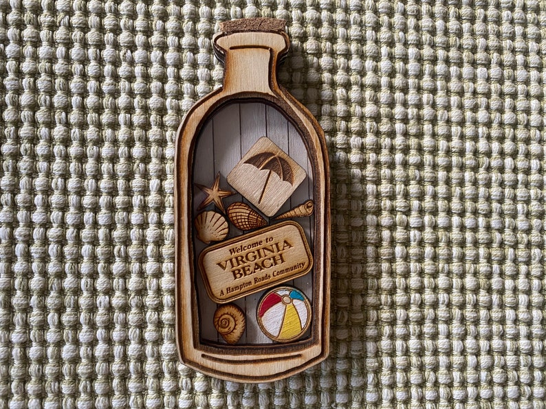 May include: Wooden bottle-shaped ornament with a cork top. Inside the bottle are various wooden cutouts including a beach umbrella, seashells, a starfish, a seashell, a beach ball, and a sign that reads "Welcome to Virginia Beach, A Hampton Roads Community".