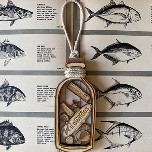May include: A wooden ornament shaped like a bottle with a cork top and a rope tied around the neck. The bottle is filled with seashells, a starfish, and a wooden sign that says "Wildwoods".