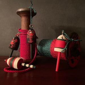 May include: A spool of red and green yarn with lobster-shaped buoys attached. The buoys are painted red and white with black accents. The spool is made of wood and has a metal handle.