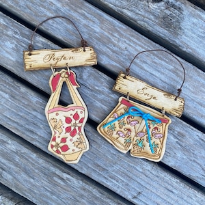May include: Two wooden ornaments with names "Peyton" and "Enzo" written on them. The ornament for Peyton has a red and white swimsuit with floral print hanging from it. The ornament for Enzo has a pair of blue and white swim trunks with pink flamingos and pineapples printed on them.