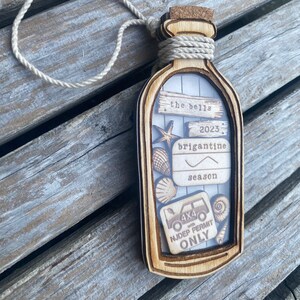 May include: A wooden ornament shaped like a bottle with a cork top. The bottle has a clear window that shows a variety of items including a seashell, a starfish, a small car, and a sign that says "the bells 2023 brigantine season 4x4 with NJDEP permit only".