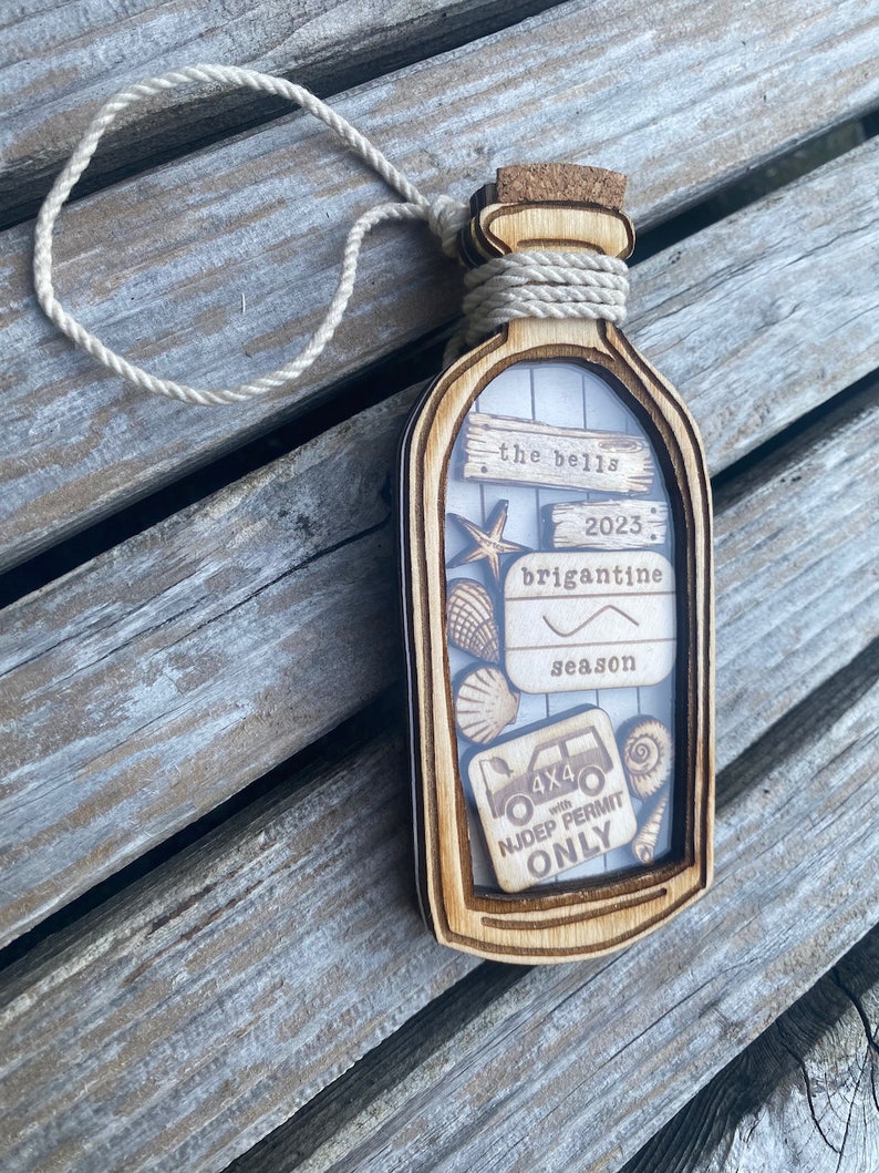 May include: A wooden ornament shaped like a bottle with a cork top. The bottle has a clear window that shows a variety of items including a seashell, a starfish, a small car, and a sign that says "the bells 2023 brigantine season 4x4 with NJDEP permit only".