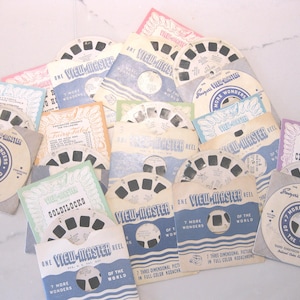 May include: A collection of vintage View-Master reels in paper sleeves, featuring titles like 7 More Wonders of the World, Cinderella, and Goldilocks. The circular reels have seven pairs of image frames, and the sleeves display various colours.